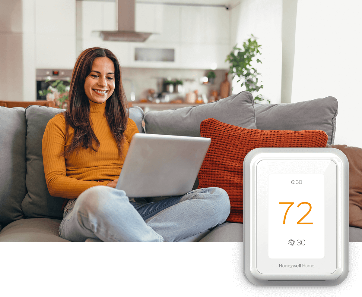A young woman relaxes with her laptop amongst pillows on a sofa in a bright living room; a smart thermostat is in the foreground