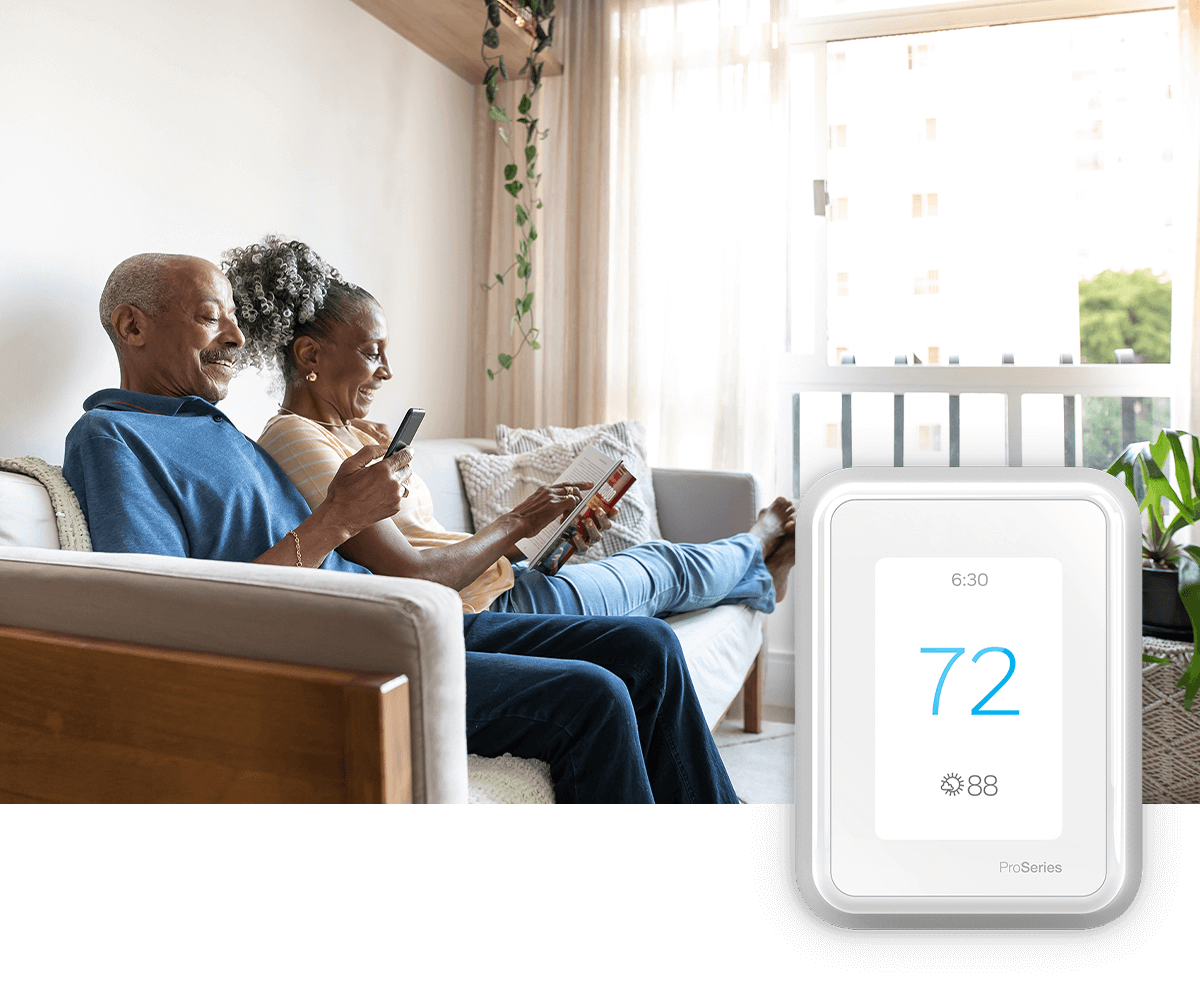 An older couple relaxes together on a sofa: one reading, one looking at a phone; a thermostat image is in the foreground