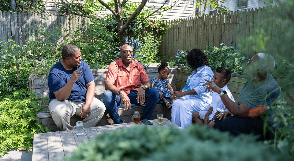 A family relaxes outside on wooden benches, enjoying the trees and conversation.