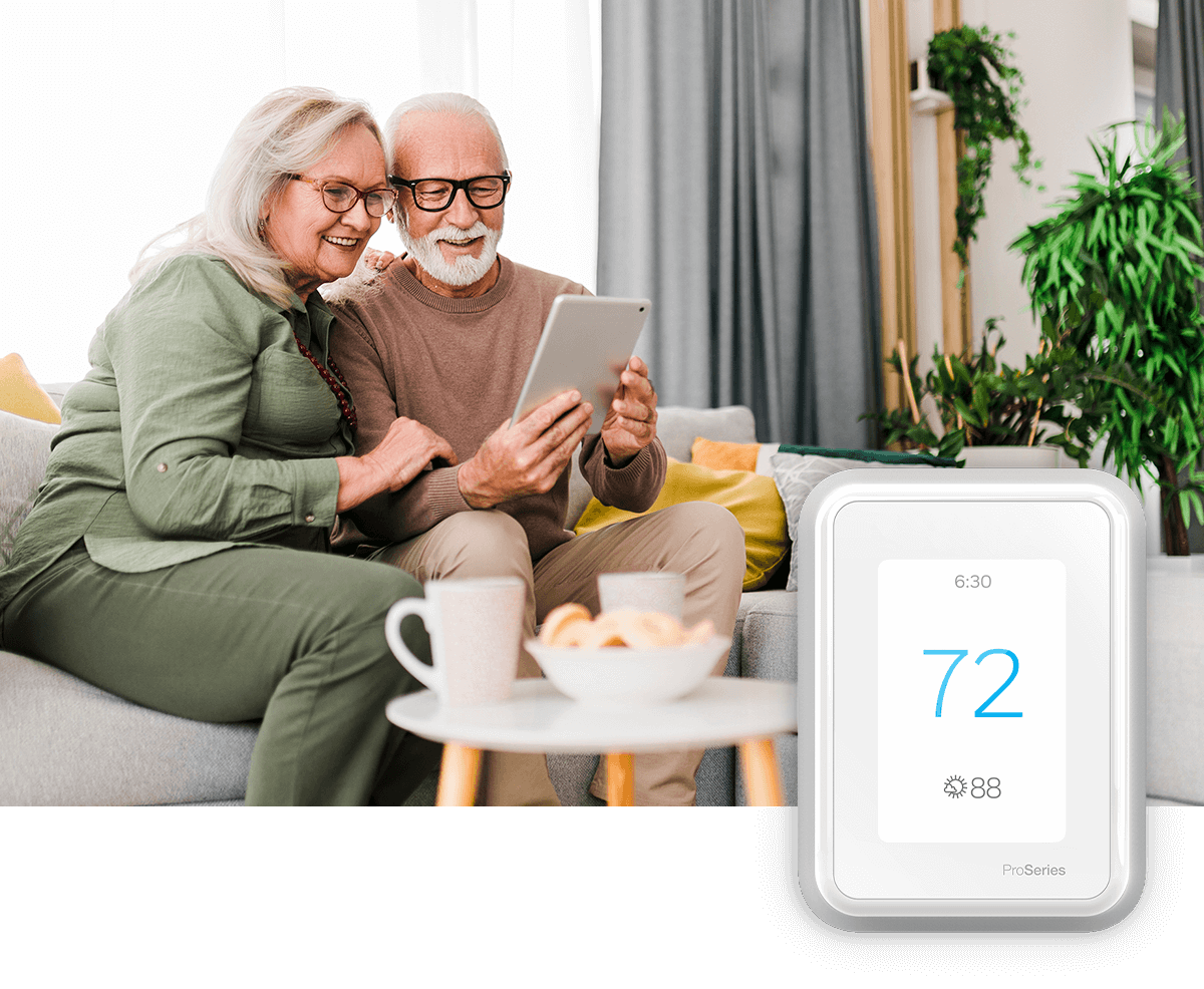 An older couple is seen smiling at a tablet while sitting on a sofa in a bright room; a thermostat image is in the foreground