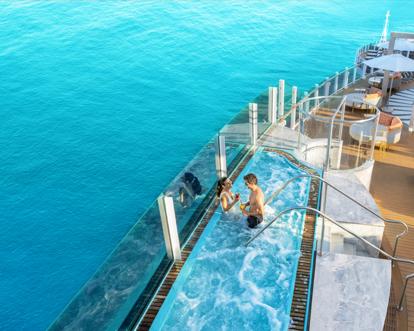 A couple relaxes in a cruise ship's infinity pool overlooking vast turquoise ocean waters. The scene exudes tranquility and luxury on a sunny day.