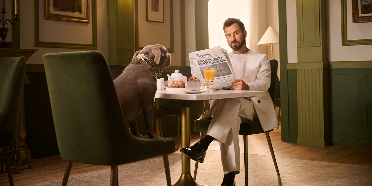 A man in a white suit reads a newspaper at a dining table, with a large dog sitting opposite.