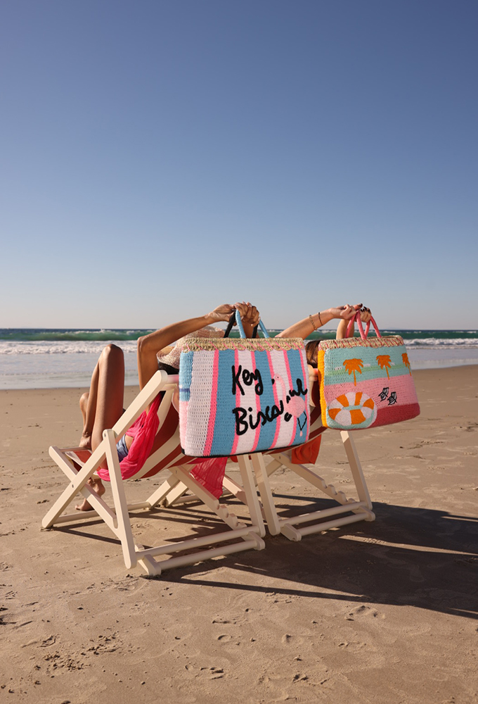 Two people relax on a sunny beach in deck chairs, their faces hidden by colorful beach bags.