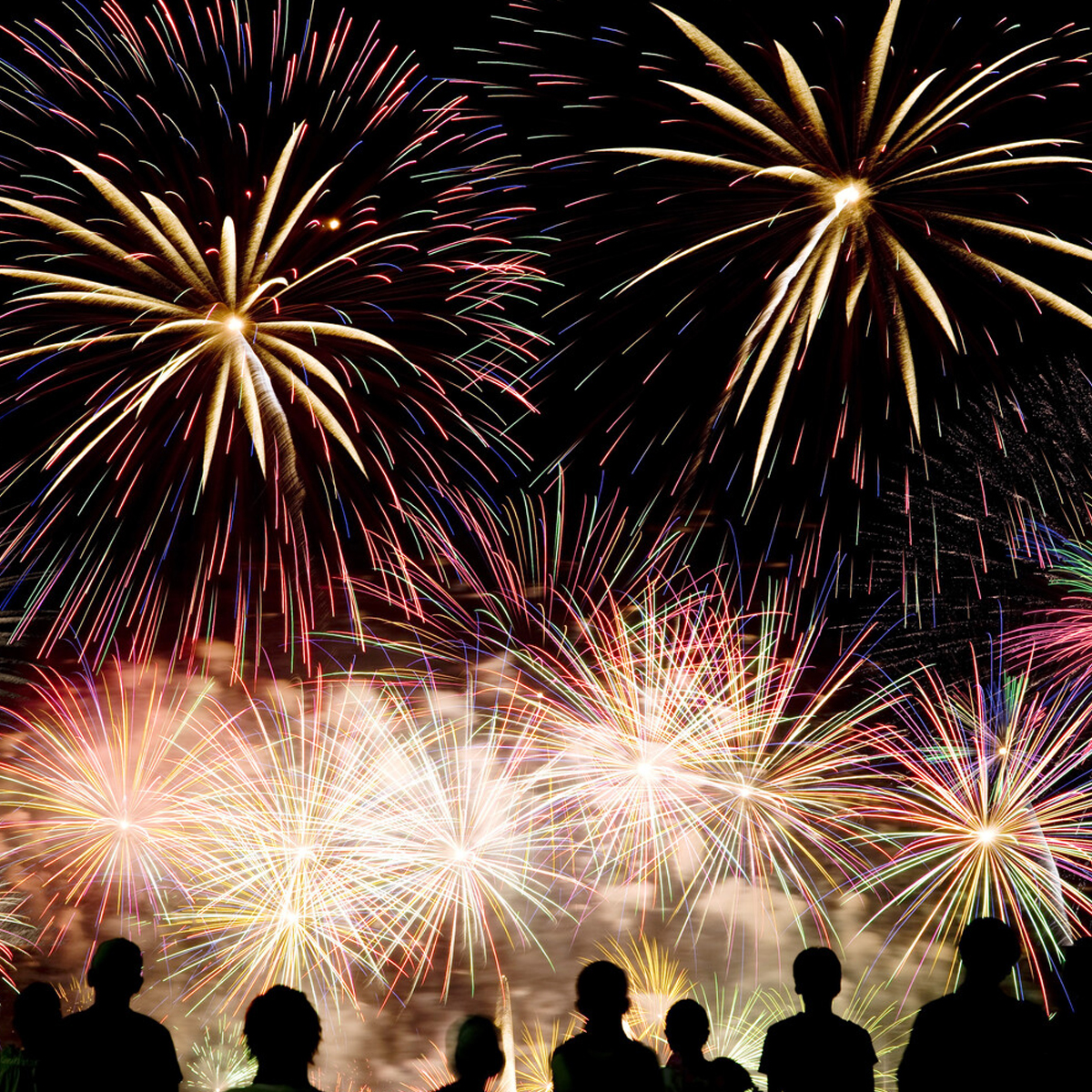 Silhouetted people watch vibrant fireworks burst across the night sky in a colorful celebration.