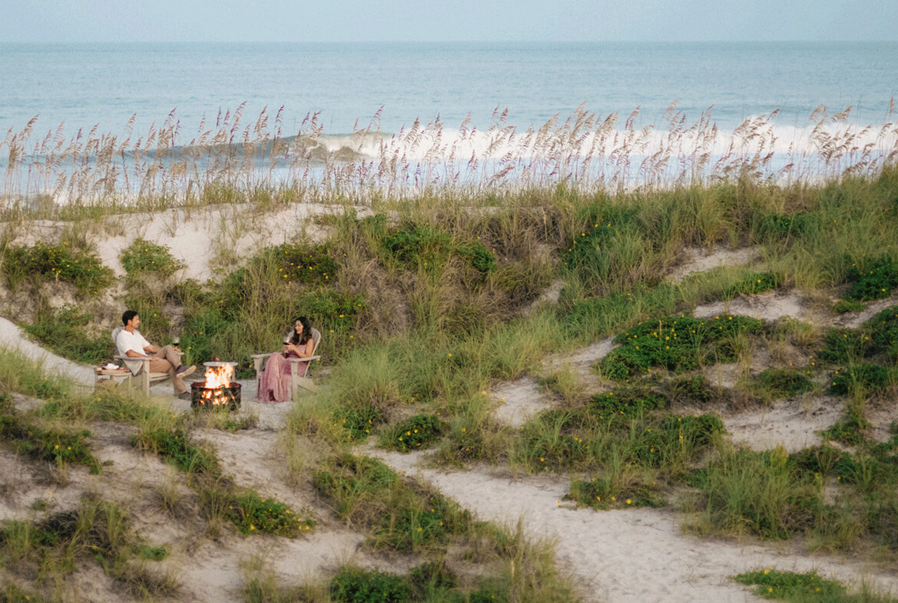 A man and woman sit by a firepit on the beach.
