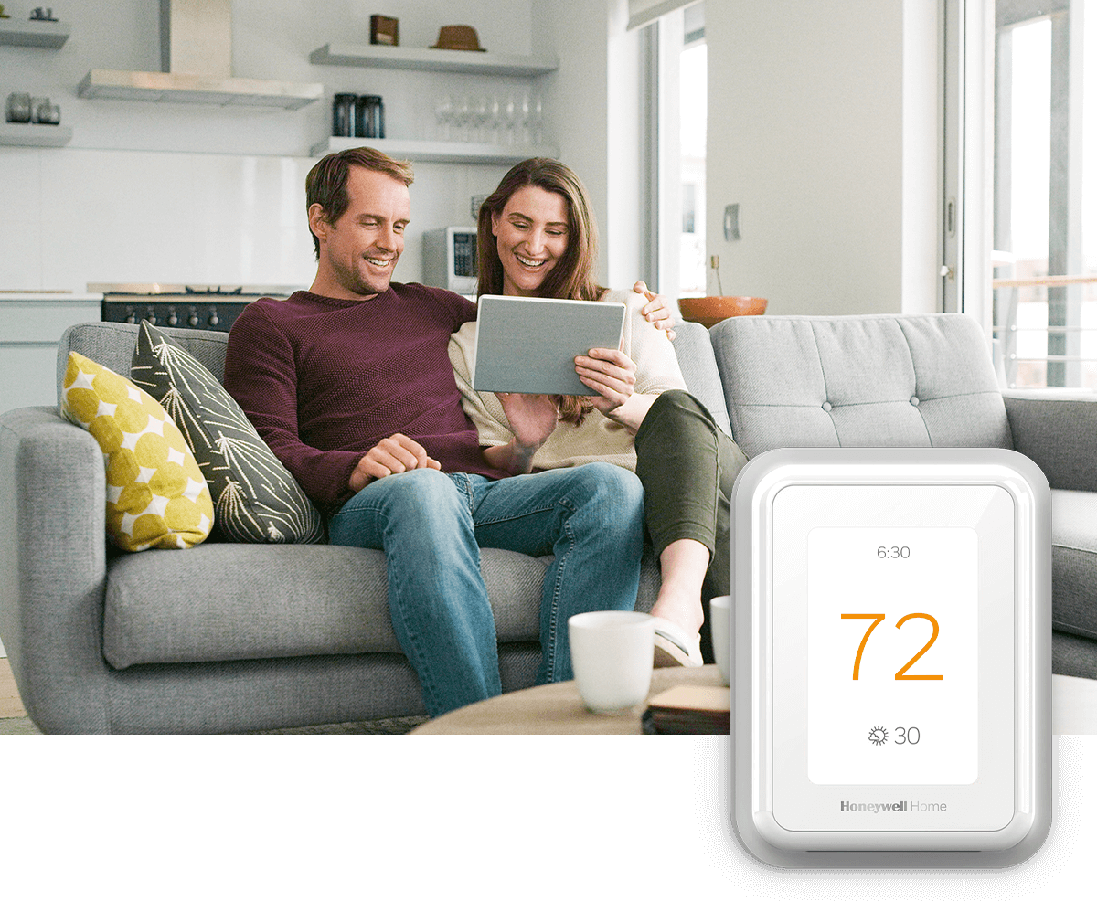 Smiling couple sitting on couch together looking at tablet; a thermostat image is in the foreground
