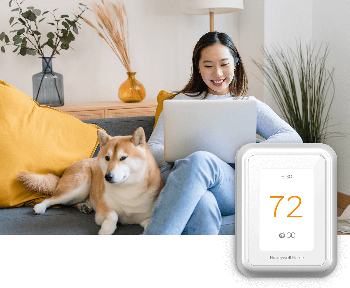 A smiling young woman sits on her couch with a dog while operating a laptop computer; a thermostat image is in the foreground