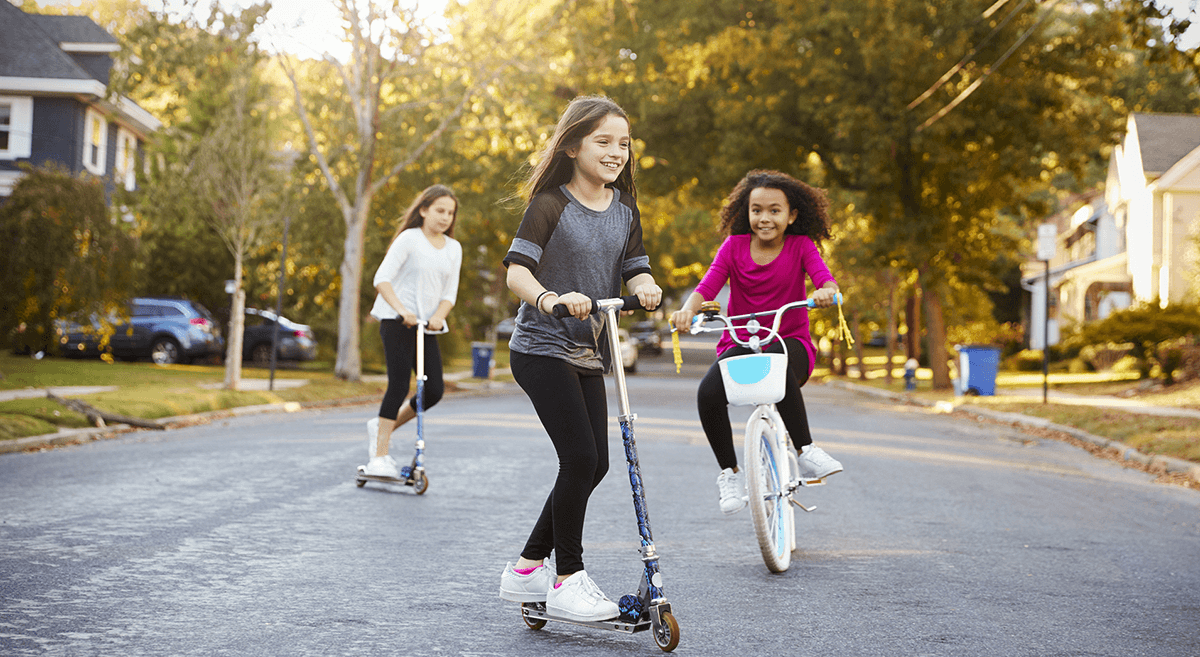 Kids ride scooters and a bicycle down a suburban street