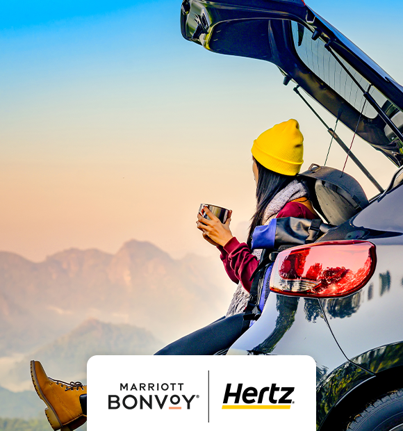 A girl wearing a yellow beanie is holding a mug while looking out from the trunk of a car.