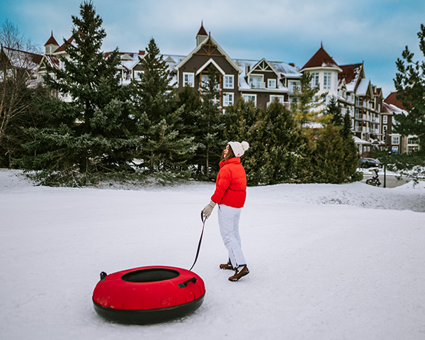A woman in a bright red jacket is going sledding in the snow in front of a ski resort.