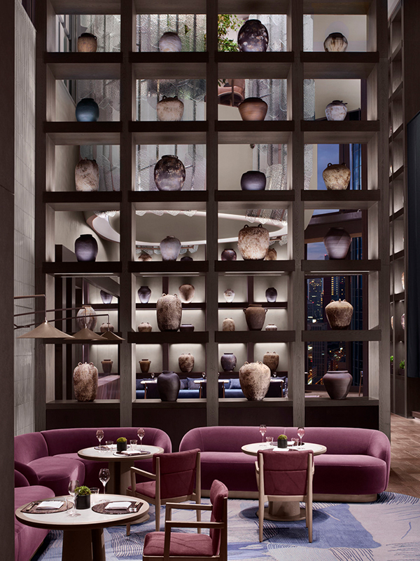 Elegant modern restaurant with purple velvet seating, wooden tables, and a towering backlit wall displaying ceramic vases.