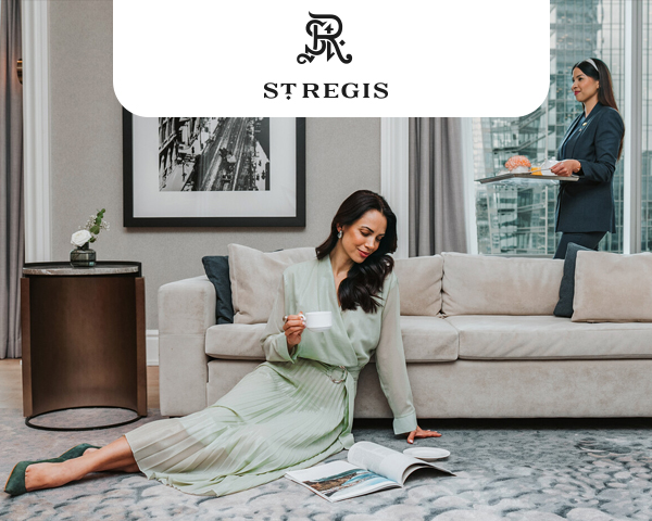 A woman in an elegant pastel green dress sits on a plush carpet in a luxurious hotel suite, holding a cup and smiling.