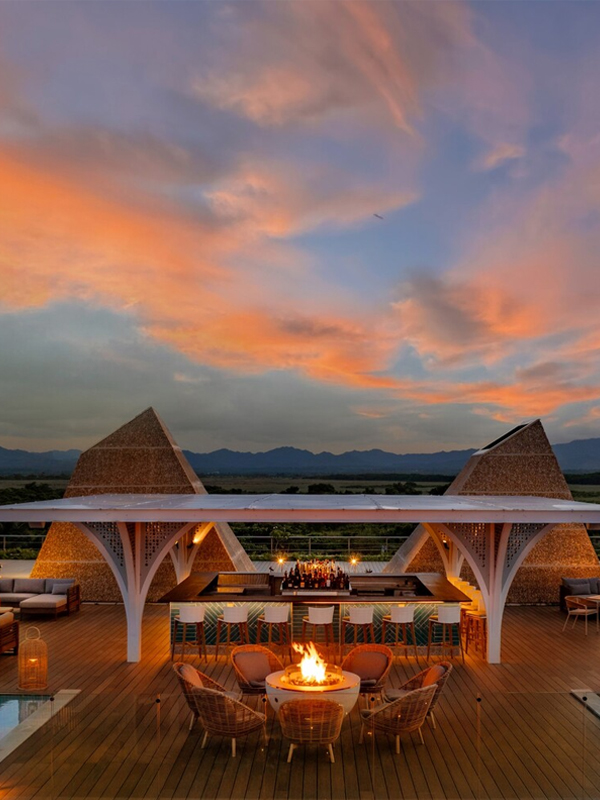 Modern architectural entrance with a circular wooden canopy supported by slender beams, lush greenery, palm trees, and vibrant tropical flowers under a clear blue sky.