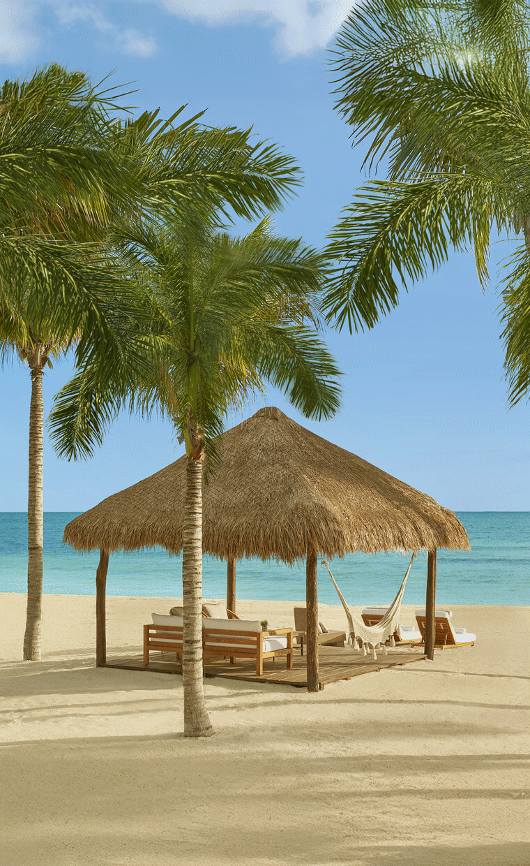 Cabana with lounge chairs on a beach with palm trees by a turquoise blue sea.
