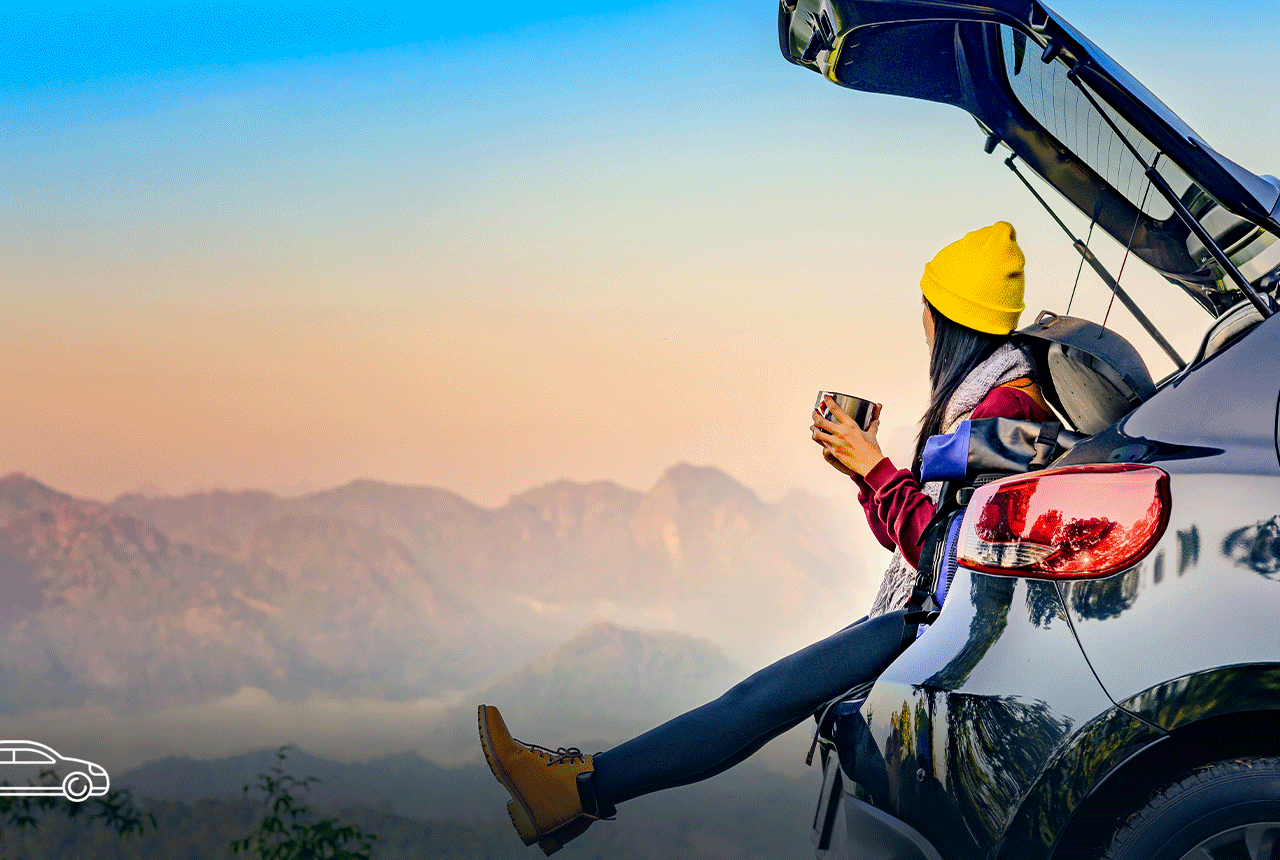 Girl wearing a yellow hat is seen sitting in the open truck of an SUV and looking out at the sunset, while also holding a mug in her hands.