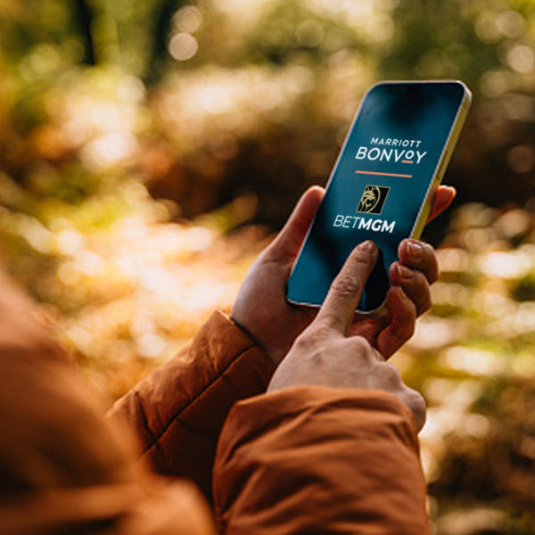 A person in an orange jacket holding a smartphone with Marriott Bonvoy and BetMGM logos on the screen, set against a blurred autumn background.