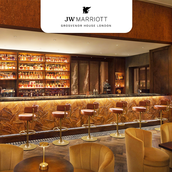 A hotel bar featuring a stocked backlit liquor shelf, barstools, and mustard-colored lounge seating.