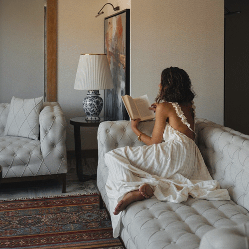 A woman in a casual white dress lounging on a couch reading a book.