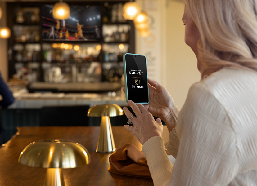 Woman in sports bar smiling at her phone. Marriott Bonvoy and BetMGM logos are visible on her phone screen.
