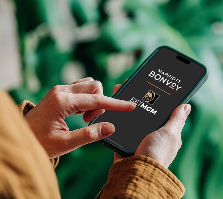A person in a brown jacket uses a smartphone with the Bet MGM and Marriott Bonvoy logos. The background is blurred, with green leaves creating a natural feel.