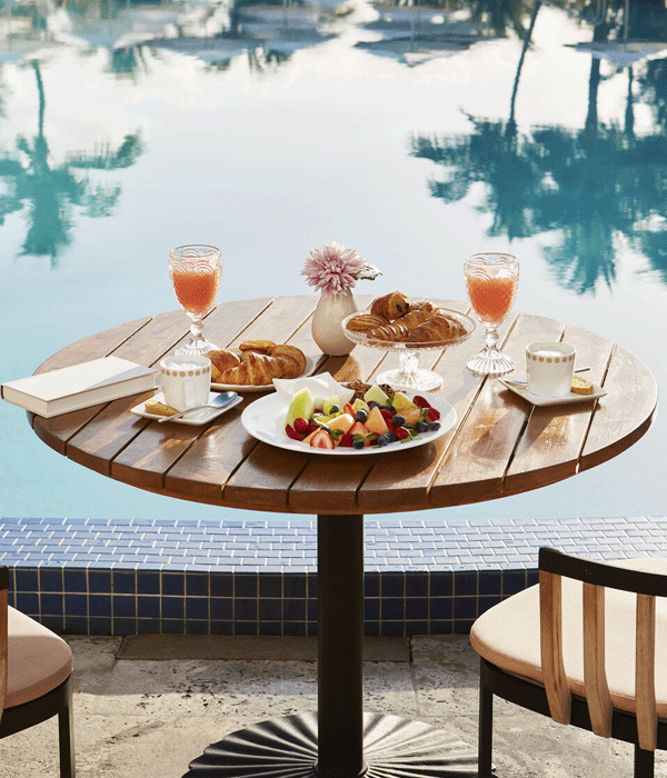A round wooden table by a pool with two chairs, set with pastries, fruit, drinks, and a small flower arrangement.