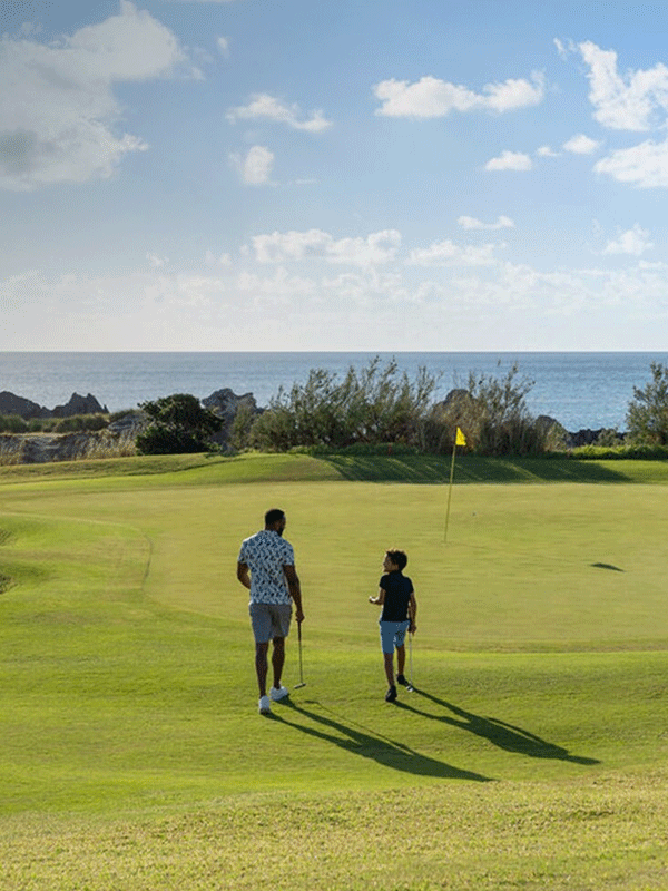 Father and son walk on a golf course near a putting green with an ocean view in the background under a partly cloudy sky.