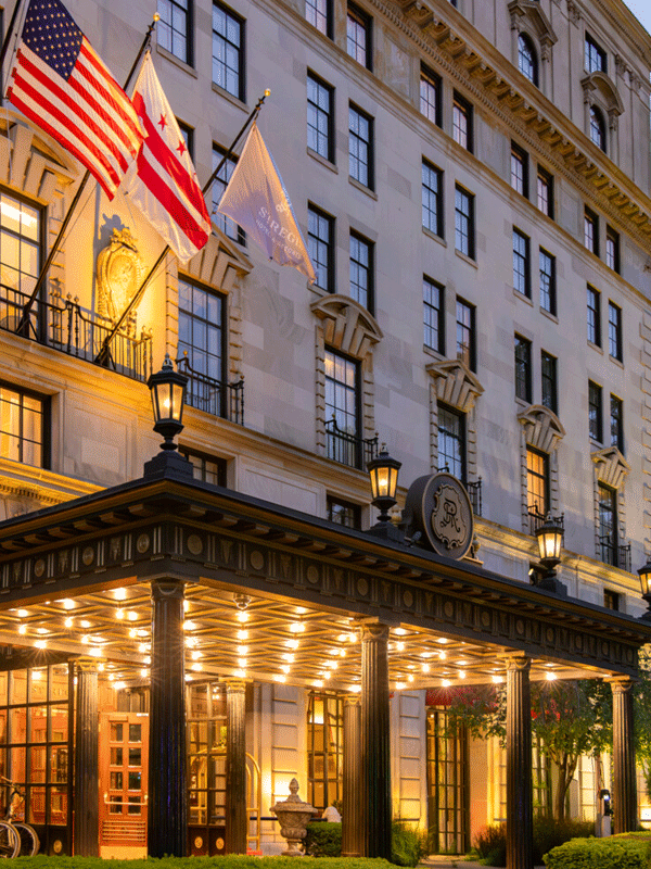 Elegant hotel entrance illuminated at dusk, displays U.S. and local flags.